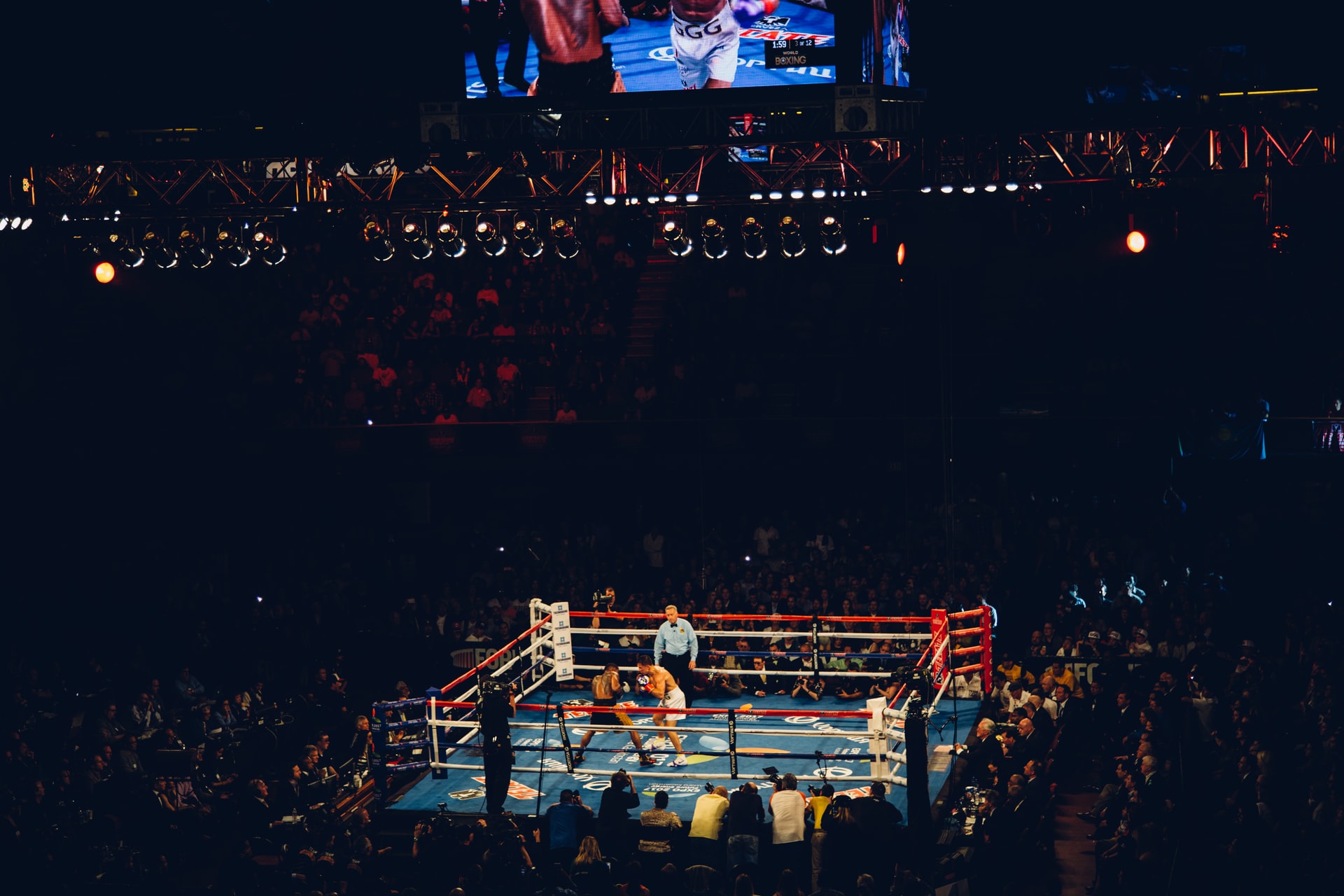 boxing arena in centre with two fighters and a referee, surrounding film crew and thousands of people in audience watching